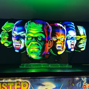 May include: A set of five colorful monster masks, each with a unique expression. The masks are made of plastic and have a glossy finish. The masks are arranged in a row on a black surface. The masks are lit from below, creating a dramatic effect.