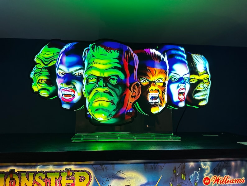 May include: A set of five colourful monster masks, each with a unique expression. The masks are made of plastic and have a glossy finish. The masks are arranged in a row on a black surface. The masks are lit from below, creating a dramatic effect.