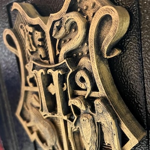 May include: A detailed, gold-colored relief of a crest, featuring a snake, eagle, badger, and lion, set against a textured black background. The crest has the letters "H" and "S" prominently displayed. The design is likely from a fantasy theme.