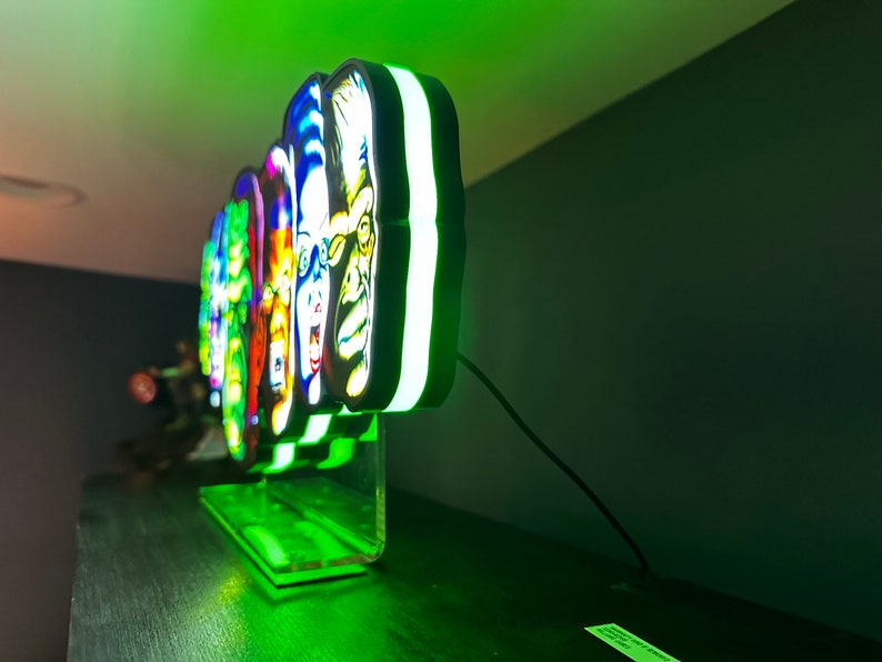 May include: A multi-coloured neon sign featuring five different faces, each with a unique expression. The sign is illuminated with green and white lights and sits on a clear acrylic stand.