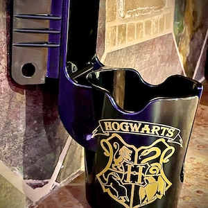 May include: A black and purple wall-mounted cup holder featuring the Hogwarts crest in gold. The holder has a bracket for mounting and a curved design to hold a cup. The Hogwarts logo is prominently displayed.