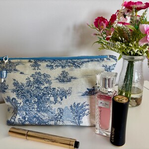 May include: A toile-patterned cosmetic bag with a blue zip and charm, next to a perfume bottle, a black lipstick tube, a mascara tube, and a vase of pink flowers. The bag features a blue and white pastoral scene.
