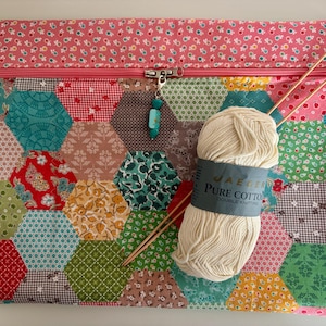 Puede incluir: Un estuche de tela rectangular con un mosaico de patrones hexagonales multicolores en rojo, verde, azul y amarillo, rematado con un borde floral rosa. Un ovillo de lana color crema y dos agujas de tejer de madera descansan sobre el estuche.