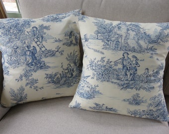 Puede incluir: Dos almohadas de color crema con un patrón de toile azul que representa una escena pastoral con figuras, árboles y una pequeña casa.