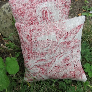 Op de afbeelding: Twee rode en witte kussens met een toile-patroon. De kussens liggen op een bed van groen gras.