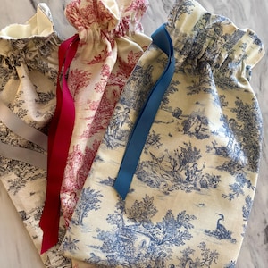 Op de afbeelding: Vier stoffen cadeauzakjes met trekkoord, met toile de Jouy-patronen in crème, rood en blauw. Elk zakje heeft een contrasterend lint. Ideaal voor kleine cadeaus of opslag, deze zakjes zijn gemaakt van stof.