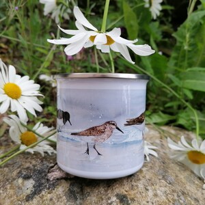 May include: White enamel camping mug featuring a watercolor illustration of shorebirds on a beach. The mug has a silver rim and a white background with a light blue and gray wash. Daisies surround the mug.