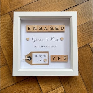 May include: A white picture frame with a white mount and a white background. The frame contains a scrabble tile arrangement that spells "ENGAGED" with two hearts and the text "Grace & Ben" and "22nd October 2021". Below the text is a brown tag with the text "The day she said..." and a heart. To the right of the tag is a scrabble tile arrangement that spells "YES".