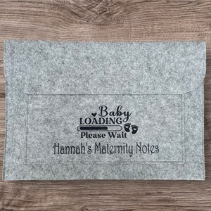 May include: Gray felt folder with a black graphic that says "Baby Loading" with a progress bar and footprints. The text "Please Wait" is below the graphic. The text "Hannah's Maternity Notes" is below the progress bar.