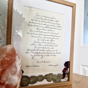 May include: A framed print featuring black calligraphy text on cream paper, enclosed in a light wood frame. The text is a quote by Louis de Bernieres, dated 18th February 2022, with the names Martin & Natasha. Dried eucalyptus leaves and dark flowers decorate the bottom.