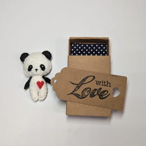 May include: A small, white and black panda bear plush toy with a red heart on its chest is in a small brown box with a blue and white polka dot fabric lining. A brown tag with the text "with Love" is attached to the box.