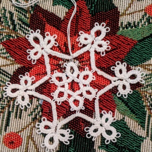 May include: A white crocheted snowflake ornament with a delicate design. The snowflake is hanging on a red and green patterned fabric.