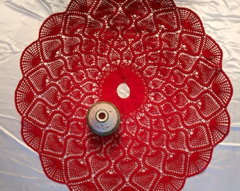May include: Red crocheted Christmas tree skirt with a white and green ceramic ornament in the center.