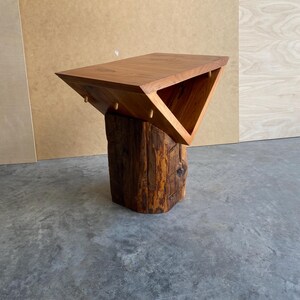 May include: A wooden table with a triangular top and a unique, rustic base made from a tree trunk. The table is made of light brown wood and has a natural, organic look.