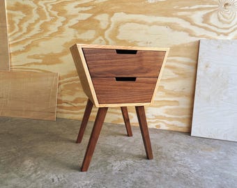 May include: A mid-century modern style nightstand with two drawers. The drawers are a dark walnut color, and the frame is a lighter wood. The nightstand has three angled legs. The overall design is minimalist and stylish.