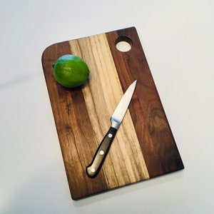 May include: A wooden cutting board with a light brown and dark brown wood grain pattern. The board has a round hole in the top right corner and a small lime on the surface. A silver knife with a black handle is lying on the board.