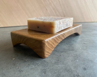 May include: A wooden soap dish with a curved design, holding a rectangular bar of soap. The dish is made of light brown wood with visible grain, and the soap has a light beige color with visible flecks.