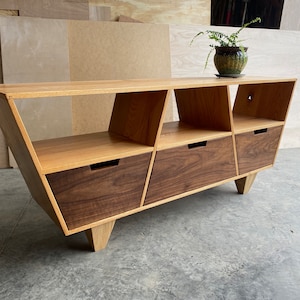 May include: A mid-century modern style wooden media console with three drawers and open shelves. The console is made of light wood with walnut drawer fronts and has angled legs.