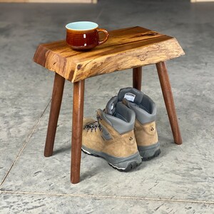 May include: A small, handcrafted wooden stool with a natural edge top and dark brown legs. A brown mug with a light blue interior sits on the stool. Hiking boots are placed beneath the stool.
