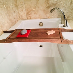 May include: A wooden bathtub tray with curved handles, holding a red book, a gray stone, and a bar of soap. The tray is made of dark wood and spans the width of a white bathtub.