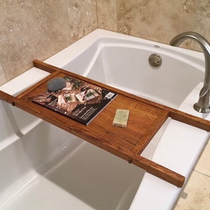 May include: A wooden bath tray with a magazine, a bar of soap, and a stone on top. The tray is in a white bathtub with a silver faucet.