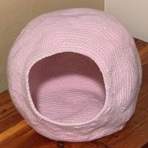 May include: A pink crocheted pet bed shaped like a dome with a round opening. The bed is made of soft yarn and has a cosy interior.