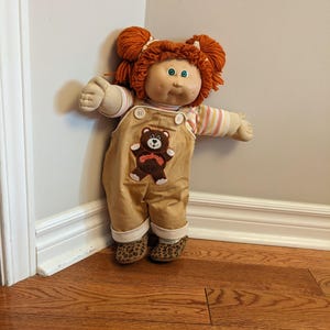 Peut inclure: Poupée Cabbage Patch Kids vintage avec des cheveux roux en deux chignons. La poupée porte une chemise rayée sous une salopette beige avec un écusson ours en peluche. Les chaussures de la poupée ont un motif léopard.