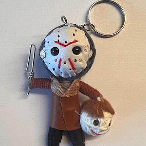 Op de afbeelding: Een handgemaakte sleutelhanger met een kleine, bruine wollen pop met een wit masker en een rode streep over het gezicht. De pop houdt een kleine, witte wollen bal met een gezicht vast. De sleutelhanger is bevestigd aan een zilveren sleutelring.