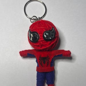 Puede incluir: Un llavero de Spider-Man hecho a mano. La cabeza es una bola de hilo rojo con detalles de ojos negros. El cuerpo es de hilo rojo y azul, con un emblema de araña negra en el pecho. El llavero tiene un anillo de metal plateado.