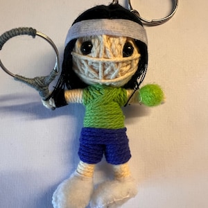 May include: A yarn doll keychain with a tennis racket and ball. The doll is wearing a green shirt, blue shorts, and a white headband. The doll has black hair and a white face.
