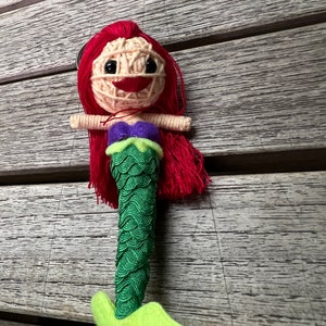 Puede incluir: Una muñeca sirena de pelo rojo con cola verde y top morado. La muñeca está hecha de hilo y tiene una cara sonriente.