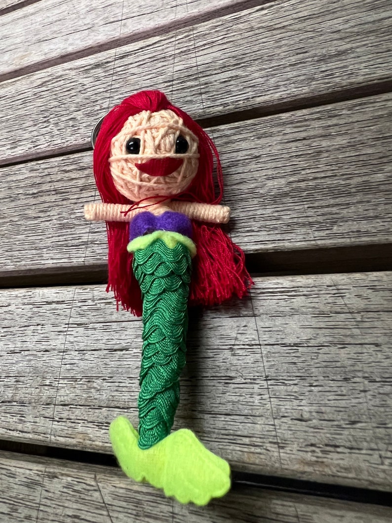 May include: A red-haired mermaid doll with a green tail and a purple top. The doll is made of yarn and has a smiling face.