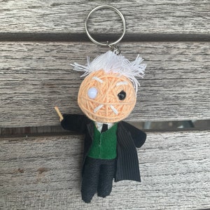 Puede incluir: Un llavero hecho a mano con una muñeca de hilo que se asemeja a un profesor. La muñeca tiene una cabeza de hilo color melocotón con pelo blanco, un traje negro, un chaleco verde y una pequeña varita de madera. El llavero tiene un anillo plateado.
