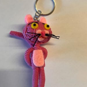 May include: Pink yarn keychain with a pink felt belly and black yarn whiskers. The keychain is shaped like a cartoon cat with large yellow eyes.
