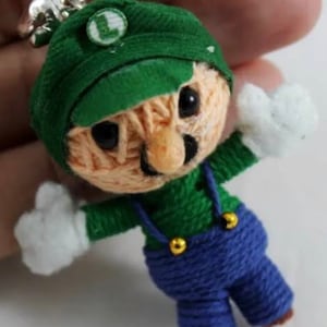Puede incluir: Un llavero de Luigi hecho a mano. Presenta un gorro verde con una "L", una camisa verde, un peto azul y guantes blancos. Tiene ojos negros y una nariz beige. Lleva un anillo plateado para llaves.