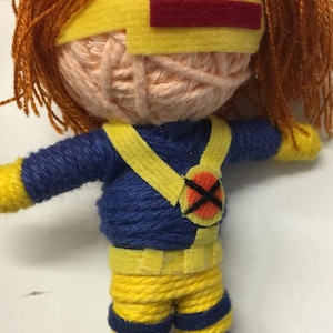 May include: A handmade yarn doll keychain featuring a character with bright orange hair, a peach-colored yarn head, and a yellow felt visor with a red rectangle. The body is wrapped in blue and yellow yarn, wearing a yellow harness with an X-logo emblem.