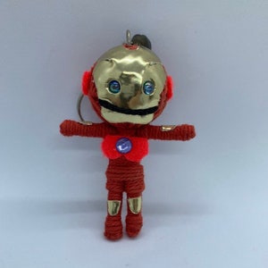 Puede incluir: Muñeca robot roja y dorada hecha a mano con un llavero. El robot tiene una cabeza metálica dorada, ojos azules y un cuerpo rojo. Los brazos y las piernas están extendidos. Una pequeña gema azul está en el pecho.