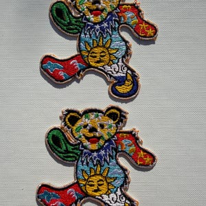 Puede incluir: Dos parches bordados idénticos que muestran un personaje de oso bailando. El oso tiene una cara amarilla con manchas verdes, un pecho azul con un gráfico de sol amarillo y extremidades con patrones rojos, azules y verdes con estrellas y nubes.