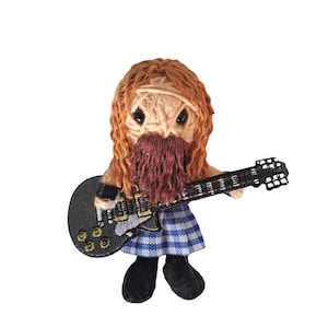 May include: A handmade, crocheted doll of a bearded musician with long red hair, wearing a blue and white plaid skirt and black shoes, holding a black electric guitar. The doll has a stern expression and is set against a white background.