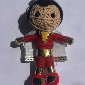 Puede incluir: Muñeca de hilo hecha a mano de un superhéroe, con un traje rojo con detalles dorados, una capa blanca y cabello negro. La muñeca tiene una cabeza envuelta en hilo con ojos negros y una campana dorada en la parte superior. La muñeca mide unos 10 cm de alto.