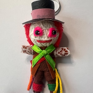 May include: A handmade doll keychain featuring a top hat, pink eyes, and red hair. The doll has a green bow tie, brown pants, and colorful yarn accents. The keychain is made of yarn, felt, and other materials.