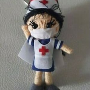 Op de afbeelding: Handgemaakte verpleegster sleutelhanger met een kleine pop met een witte muts, masker en uniform met een rood kruis. De pop heeft zwart haar en houdt een arm omhoog. De sleutelhanger is gemaakt van garen en heeft een zilveren sleutelring.