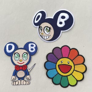 May include: Three embroidered patches including a dark blue mouse-like character head with the letters O and B, a full-body blue mouse-like character with a red bow tie and letters O and B, and a multi-colored flower with a yellow smiling face.