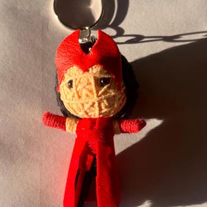 Puede incluir: Un llavero hecho a mano con una muñeca que se asemeja a un personaje con un disfraz y un tocado rojos. La cara de la muñeca está hecha de hilo beige, con cabello negro y brazos rojos. El llavero tiene un anillo plateado.