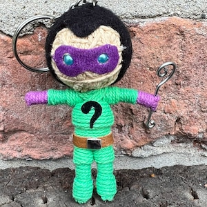 Puede incluir: Llavero artesanal del Enigma. El cuerpo es verde con un signo de interrogación negro, brazos morados y cinturón marrón. La cabeza es negra con cara beige, máscara morada y ojos azules. Incluye un llavero plateado y un signo de interrogación metálico.