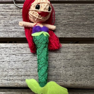 May include: A red-haired mermaid doll with a green tail and a purple top. The doll is made of yarn and has a key ring attached.
