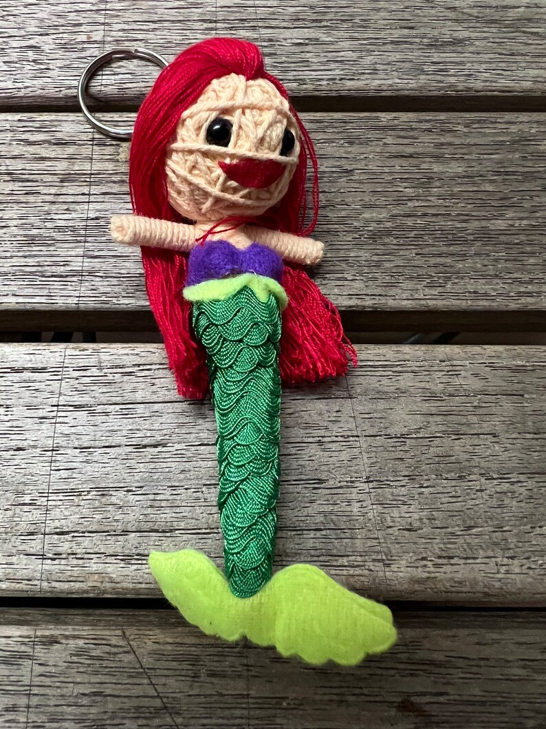 May include: A red-haired mermaid doll with a green tail and a purple top. The doll is made of yarn and has a key ring attached.