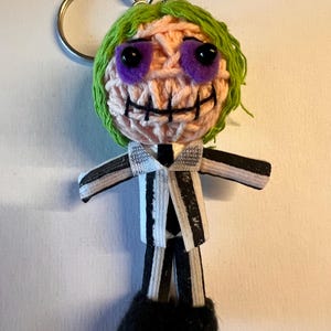 Puede incluir: Llavero hecho a mano con un personaje tejido a ganchillo con pelo verde, ojos morados y una sonrisa cosida. El personaje lleva un traje a rayas blanco y negro y zapatos negros. Un llavero plateado está adjunto.