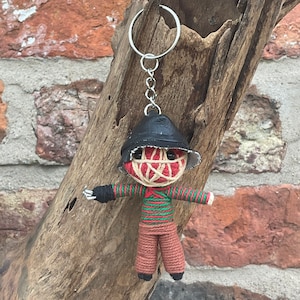 Op de afbeelding: Een handgemaakte sleutelhanger van een poppenfiguur van touw met een rood-wit gestreept masker, een zwarte hoed en een groen-wit gestreept shirt. De pop draagt een bruine broek en heeft een zwart-wit gezicht.