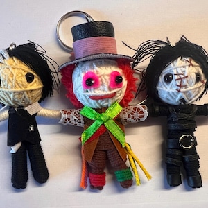 May include: Three handmade yarn doll keychains. The doll on the left is wearing a black suit and holding a knife. The doll in the middle is wearing a brown and green outfit with a pink and red hat. The doll on the right is wearing a black outfit with a white face and a stitched mouth.